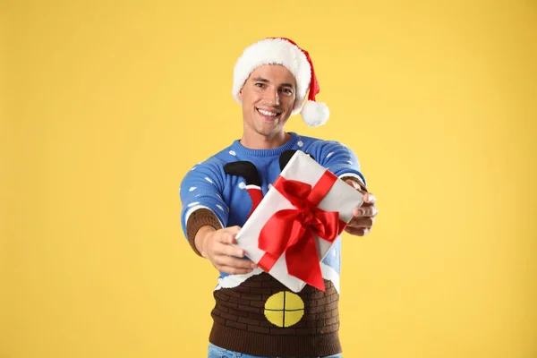 Happy man in Christmas sweater and Santa hat holding gift box on yellow background — Stock Photo, Image