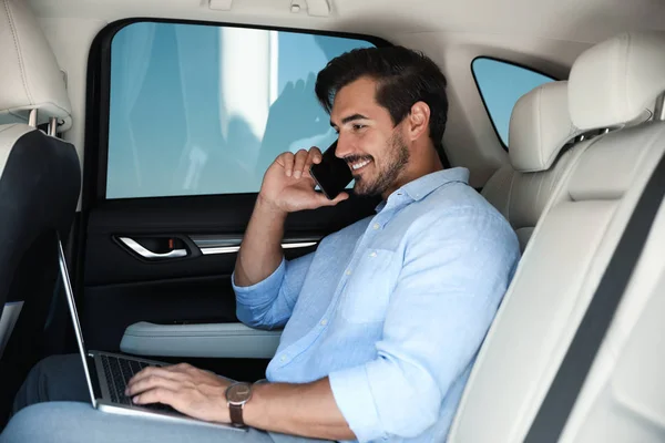 Atractivo joven que trabaja con el ordenador portátil y habla por teléfono en un coche de lujo — Foto de Stock
