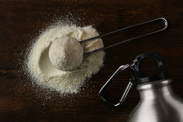 Measuring scoop of protein powder and bottle on wooden table, flat lay