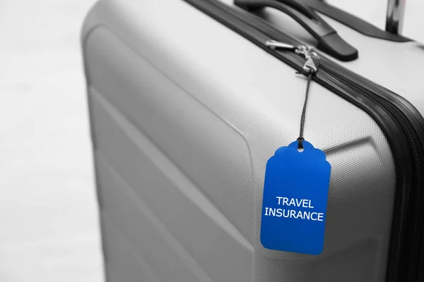 Grey suitcase with TRAVEL INSURANCE label on blurred background, closeup