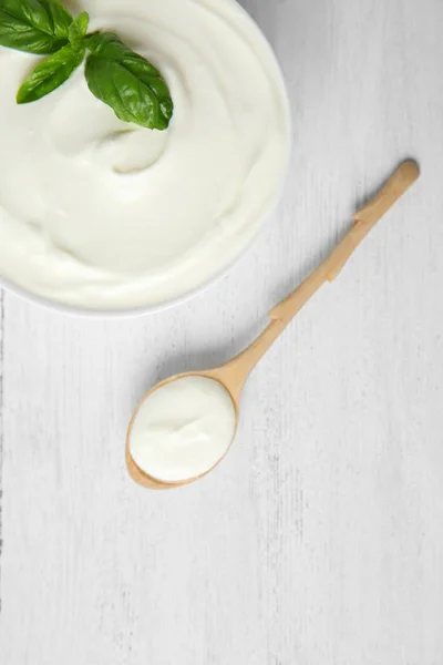 Bowl of fresh sour cream with basil and spoon on white wooden table, flat lay — Stock Photo, Image
