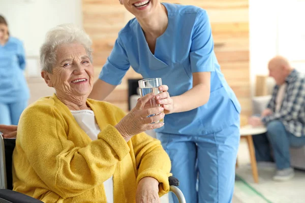 Enfermeira dando copo de água para a mulher idosa em cadeira de rodas em casa de repouso. Assistência aos idosos — Fotografia de Stock