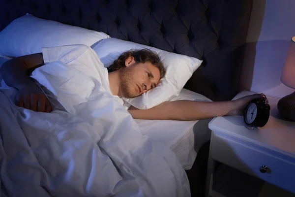 Сонный молодой человек выключает будильник на тумбочке. Время спать — стоковое фото
