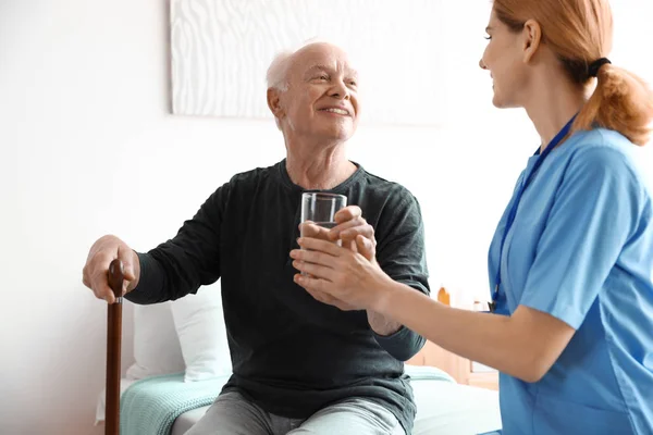 Медсестра дает стакан воды пожилому человеку в помещении. Медицинская помощь — стоковое фото