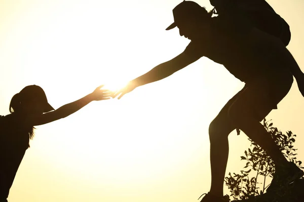 Silhuetas de homem e mulher ajudando uns aos outros a subir na colina contra o pôr do sol — Fotografia de Stock