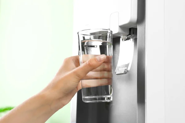 Woman filling glass from water cooler indoors, closeup. Refreshing drink
