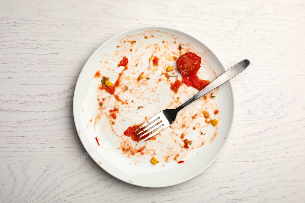 Dirty plate with food leftovers and fork on white wooden background, top view
