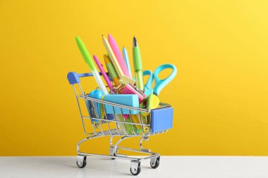 Sarı arka plan karşı beyaz ahşap masa üzerinde farklı okul kırtasiye ile küçük alışveriş sepeti