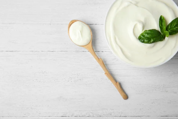 Bowl of fresh sour cream with basil and spoon on white wooden table, flat lay. Space for text — Stock Photo, Image