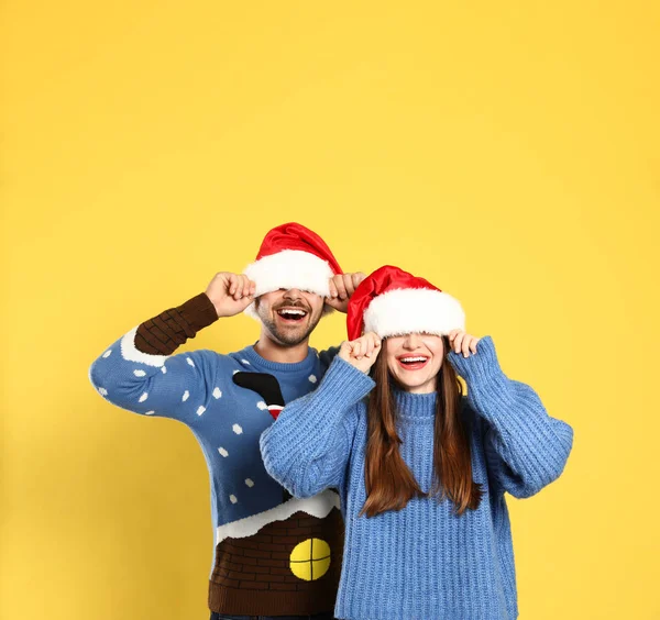 Casal usando camisolas de Natal e chapéus de Papai Noel no fundo amarelo — Fotografia de Stock