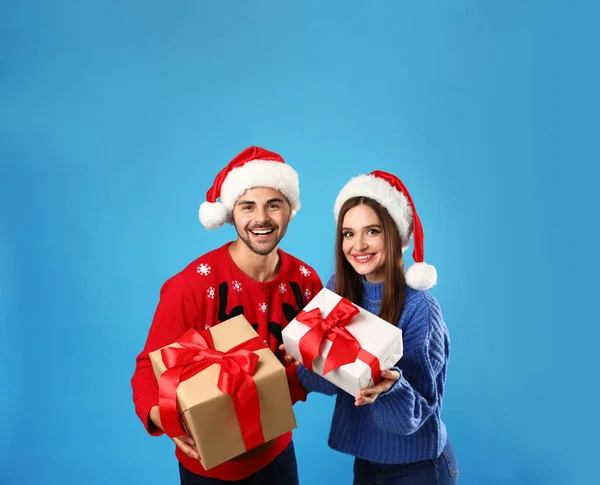 Casal em camisolas de Natal com chapéus de Papai Noel e caixas de presente no fundo azul — Fotografia de Stock