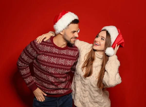 Couple portant des pulls de Noël et des chapeaux de Père Noël sur fond rouge — Photo