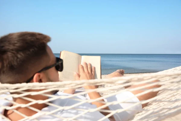 Mladý muž čte knihu v houpací síti na pláži — Stock fotografie