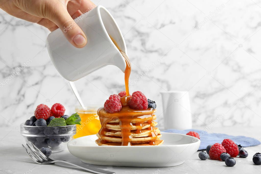 Woman pouring syrup onto fresh pancakes with berries at light grey marble table, closeup