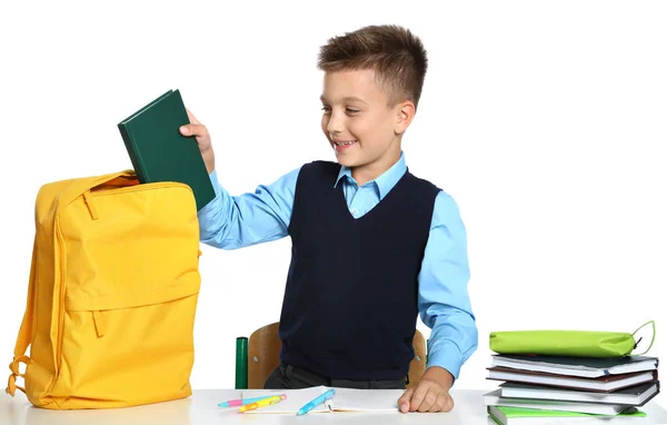 Niño en uniforme poniendo libro en la mochila en el escritorio sobre fondo blanco. Papelería escolar —  Fotos de Stock