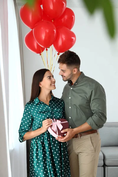 Casal jovem com balões de ar e caixa de presente em casa. Celebração do Dia de São Valentim — Fotografia de Stock