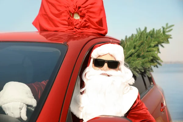 Autentico Babbo Natale con abete e borsa piena di regali sul tetto guida auto moderna vicino al mare — Foto Stock