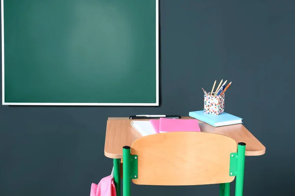 Bureau scolaire en bois avec papeterie et sac à dos près du tableau noir sur mur gris — Photo