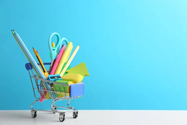 Pequeno carrinho de compras com diferentes artigos de papelaria escolar na mesa branca contra fundo azul claro. Espaço para texto — Fotografia de Stock