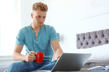 Evde yatakta otururken dizüstü bilgisayar kullanan genç adam