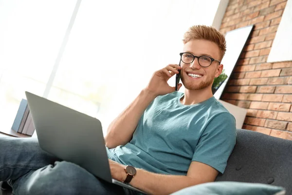 Młody człowiek rozmawia na telefon podczas korzystania z laptopa w salonie — Zdjęcie stockowe