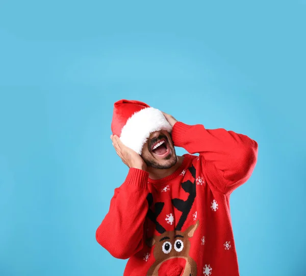 Emocjonalny młody człowiek w świątecznym swetrze i kapeluszu Świętego Mikołaja na jasnym niebieskim tle — Zdjęcie stockowe