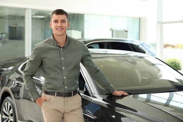 Jonge man in de buurt van nieuwe auto in moderne autodealer. Ruimte voor tekst — Stockfoto