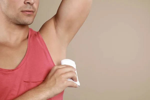 Young man applying deodorant to armpit on beige background, closeup. Space for text