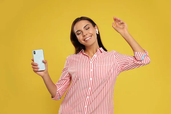 Χαρούμενη νεαρή γυναίκα με smartphone ακούγοντας μουσική μέσω ασύρματων ακουστικών σε κίτρινο φόντο — Φωτογραφία Αρχείου