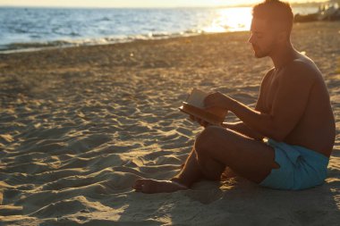 Genç adam deniz kenarındaki kumsalda kitap okuyor.