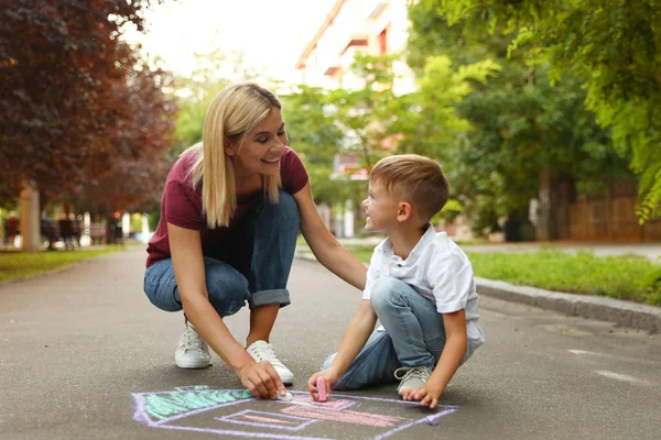 Nanny with cute little boy drawing house with chalks on asphalt — Stock Photo, Image