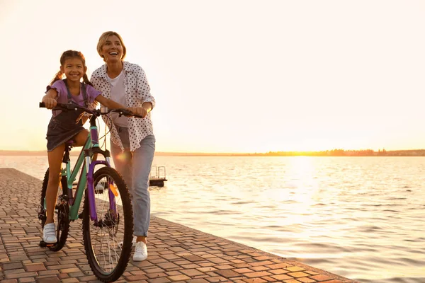 Mère heureuse enseignant à sa fille à faire du vélo près de la rivière au coucher du soleil — Photo