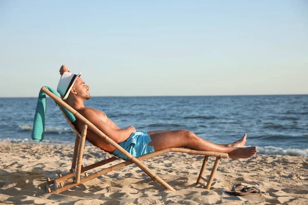 Hombre joven relajándose en la silla de cubierta en la playa cerca del mar — Foto de Stock