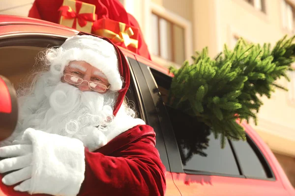 Autêntico Papai Noel com abeto e saco cheio de presentes no telhado dirigindo carro moderno, ao ar livre — Fotografia de Stock