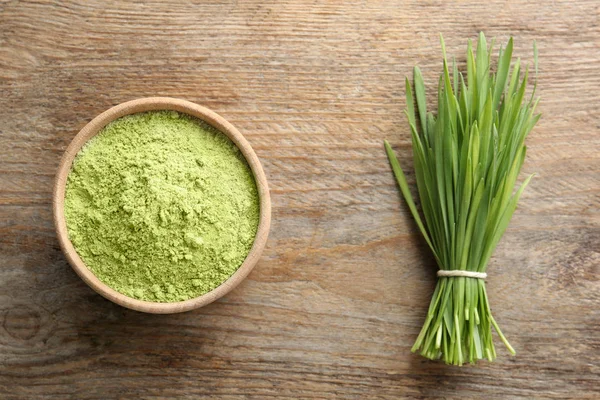 Bowl of wheat grass powder and green sprouts on wooden table, flat lay