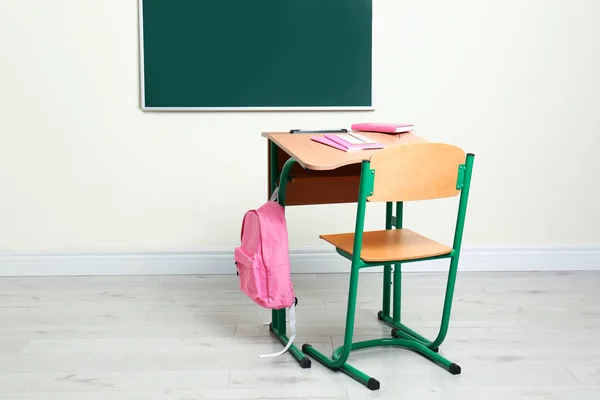 Деревянный школьный стол с канцелярскими принадлежностями и рюкзаком возле доски в классе — стоковое фото