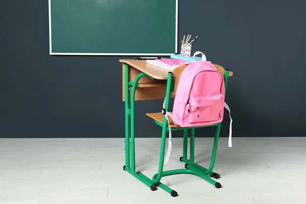 Деревянный школьный стол с канцелярскими принадлежностями и рюкзаком возле доски на серой стене — стоковое фото