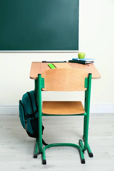 Деревянный школьный стол с канцелярскими принадлежностями, яблоком и рюкзаком возле доски в классе — стоковое фото