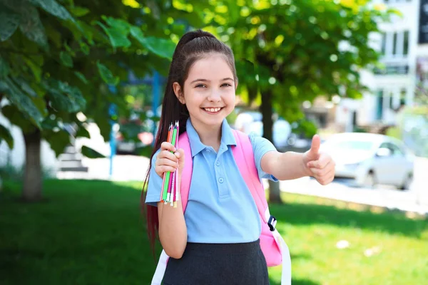 Schattig klein meisje in school uniform met rugzak en briefpapier tonen thumbs-up op straat — Stockfoto