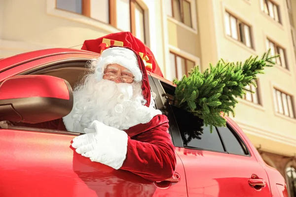 Autentyczny Święty Mikołaj z jodełką i torbą pełną prezentów na dachu jazdy nowoczesny samochód, na zewnątrz — Zdjęcie stockowe