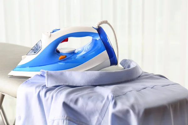 Modern electric iron and clean shirt on board against light background