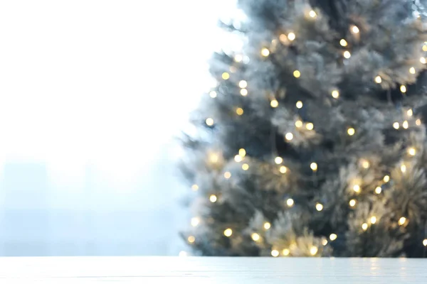 Vista borrosa de hermoso árbol de Navidad con luces amarillas cerca de la ventana interior, se centran en la mesa de madera. Espacio para texto — Foto de Stock