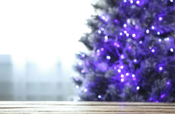 Θολή θέα του όμορφου χριστουγεννιάτικου δέντρου με μωβ φώτα κοντά στο παράθυρο σε εσωτερικούς χώρους, εστίαση στο ξύλινο τραπέζι. Χώρος για κείμενο — Φωτογραφία Αρχείου
