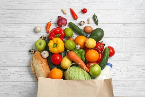 Paper bag with different groceries on white wooden table, flat lay — Stock Photo, Image