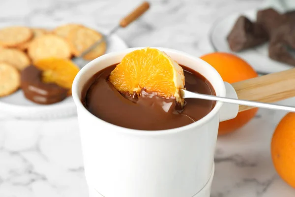 Orange in Fonduetopf mit Schokolade auf weißem Marmortisch tauchen, Nahaufnahme — Stockfoto