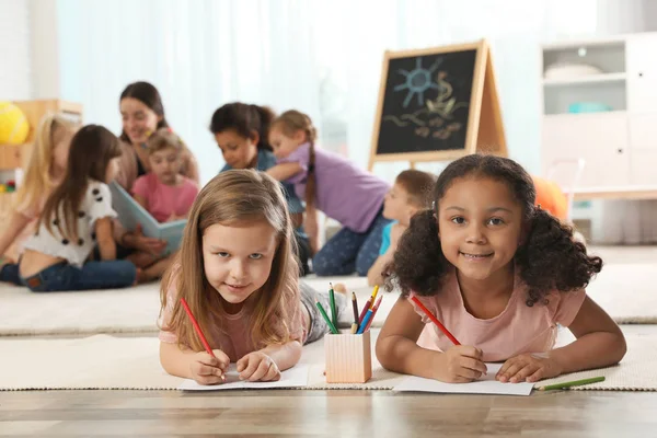 Cute girls drawing on floor while kindergarten teacher reading book to other children indoors — Stock Photo, Image