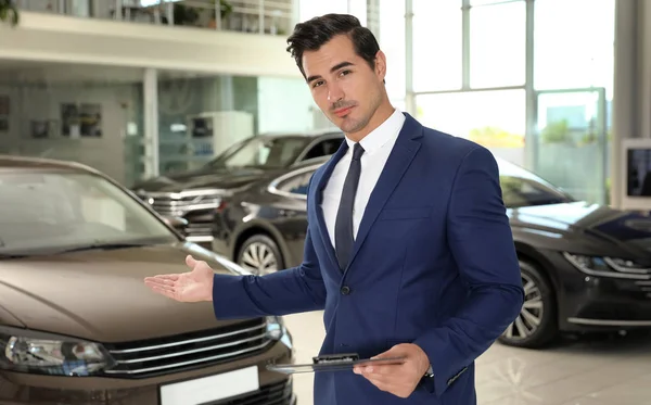 Joven vendedor con portapapeles cerca de coche en concesionario moderno — Foto de Stock