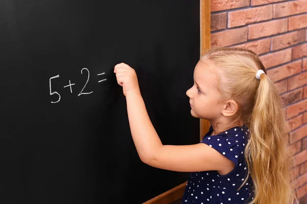 Cute little left-handed girl doing sums on chalkboard near brick wall — Stock Photo, Image