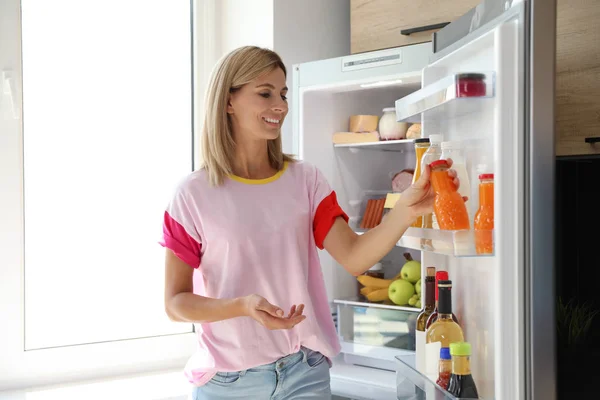 Женщина с бутылкой сока рядом с открытым холодильником на кухне — стоковое фото