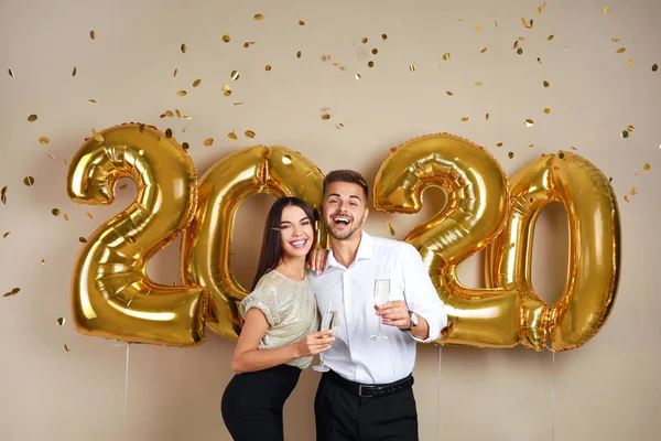 Feliz pareja joven con copas de champán cerca de globos dorados 2020 sobre fondo beige. Celebración de Año Nuevo — Foto de Stock